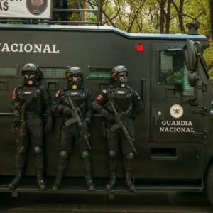 Guardia Nacional officers in tactical gear stand alert beside an armored vehicle.